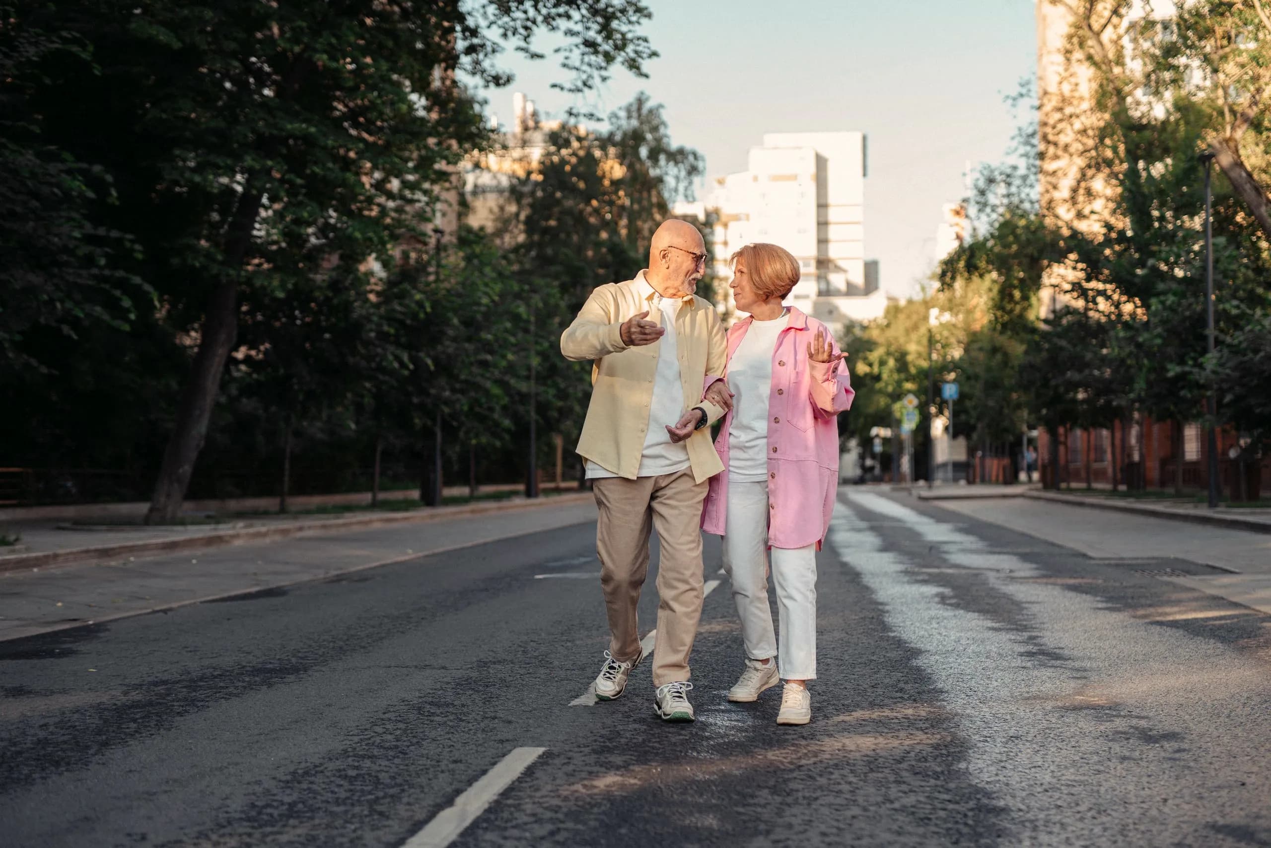 An older couple walking together along a quiet tree-lined street in warm evening light.
